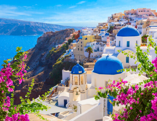 A photograph of the Greek island of Santorini in spring, with bouginvillea blooms and the blue domes of Oia in the background. The colors of the Murder Mystery Quilt for 2024 are drawn from the shades of blue, the ocean, and the pops of pink from the flowers.